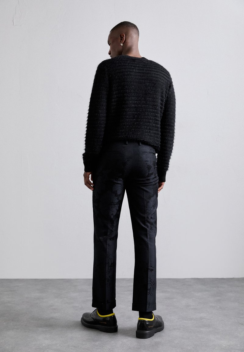 Sort strikket sweater med en cropped design, parret med mørke, mønstrede bukser. Fodtøj har sorte sko med gule accenter.