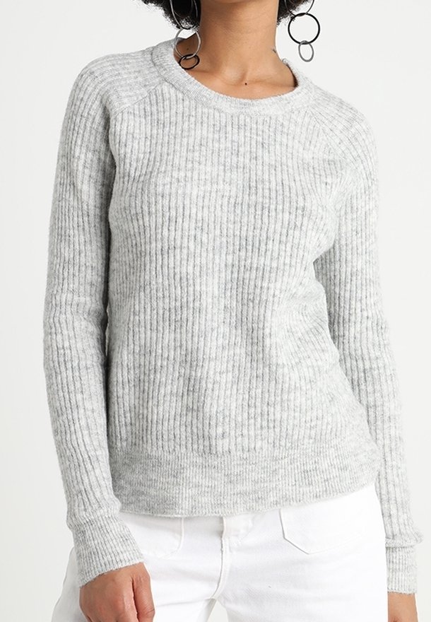 Femme portant un pull en tricot gris clair à côtes avec des manches longues, associé à un pantalon blanc, montrant le haut du torse et la main.