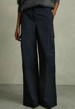 Wide-leg black cargo pants with side flap pocket, worn with gray long-sleeve sweater, hands in pockets, standing on gray floor.