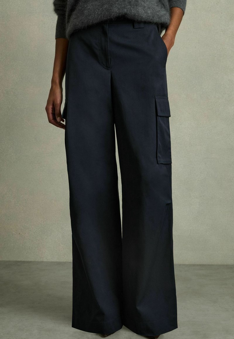 Pantalon cargo noir à jambes larges avec poche à rabat latérale, porté avec un pull gris à manches longues, mains dans les poches, debout sur un sol gris.