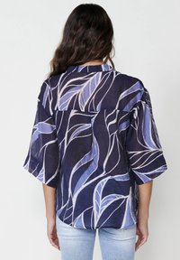 Camicia blu navy con un design fluente, caratterizzata da motivi a foglia bianchi e blu chiaro. Maniche corte e vestibilità rilassata, tessuto strutturato.