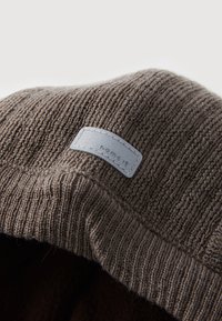 Brun strikket lue med ribbestruktur. Har et lys grått skinnmerke med preget tekst. Mykt stoff, uformell design.