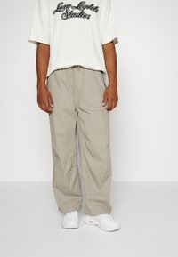 Pantaloni di cotone beige con una vestibilità rilassata e gamba affusolata, dotati di una cintura elastica e tasche laterali, abbinati a una maglietta bianca con grafica.