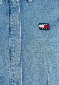 Džínsová košeľa svetlomodrej farby s gombíkovým zapínaním, ktorá na ľavej hrudi obsahuje nášivku s logom "TOMMY JEANS" v červenej, bielej a námorníckej modrej farbe.