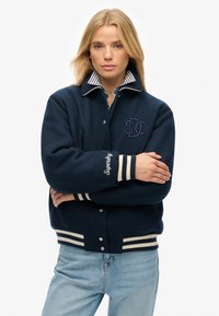 Chaqueta varsity de lana navy con cuello rayado, acentos blancos, logotipo bordado en el pecho y en el puño, y cierre de botones a presión.