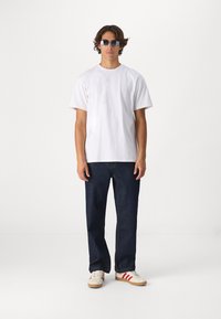 T-shirt en coton blanc, coupe ample, présenté avec un jean en denim bleu foncé. Chaussures : baskets crème avec des rayures rouges. Accessoires : lunettes de soleil bleues.