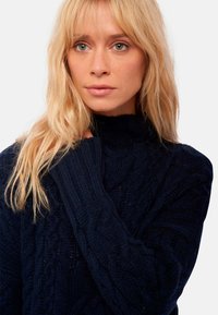 Pull en maille torsadée bleu marine avec un col montant. Présente des motifs texturés et des poignets côtelés. Fabriqué dans un matériau épais et confortable.