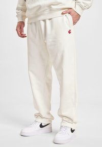 Lysbeige sweatpants med en afslappet pasform, udstyret med en diskret rød logo og sidelommer, parret med hvide sneakers med sorte detaljer.