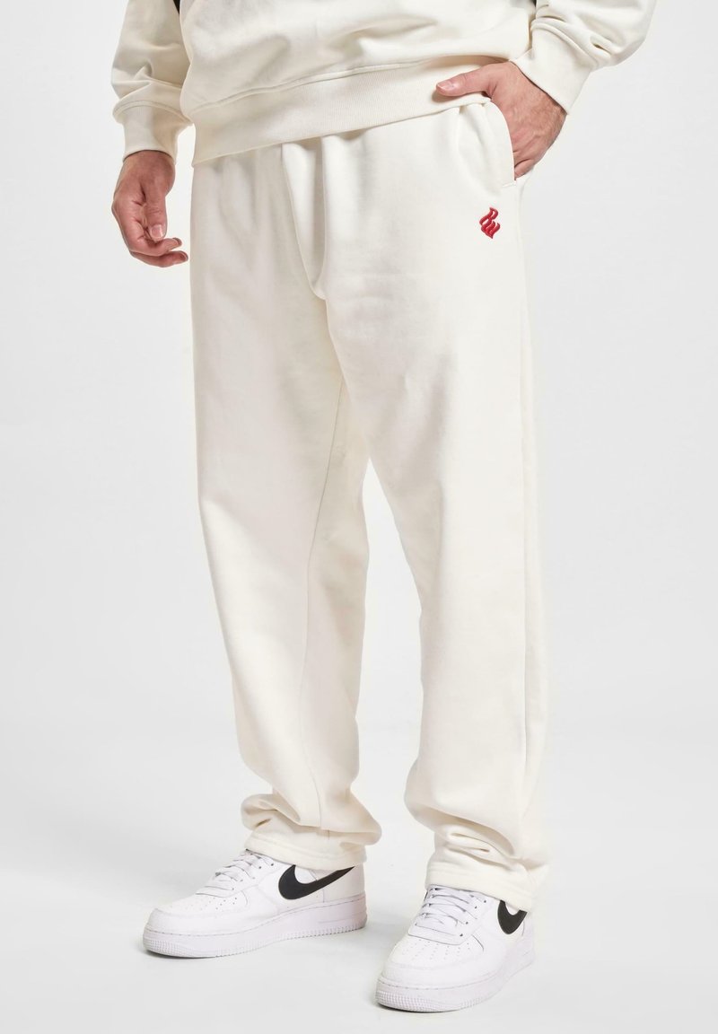 Lysbeige sweatpants med en afslappet pasform, udstyret med en diskret rød logo og sidelommer, parret med hvide sneakers med sorte detaljer.