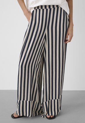 Woman wearing wide-leg pants with vertical navy and beige stripes and black sandals, standing with one hand in pocket.