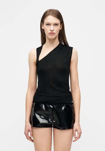 Mujer vestida con un top negro asimétrico sin mangas y pantalones cortos de cuero negro brillante, de pie contra un fondo claro y liso.
