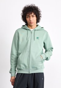 Fresca hoodie verde claro com fecho éclair, dois bolsos frontais, punhos canelados e um logo bordado da Adidas no lado esquerdo do peito. Textura de tecido suave.