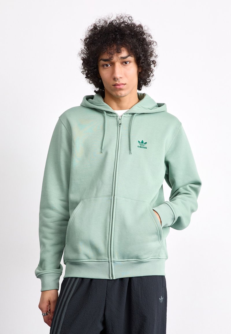 Fresca hoodie verde claro com fecho éclair, dois bolsos frontais, punhos canelados e um logo bordado da Adidas no lado esquerdo do peito. Textura de tecido suave.