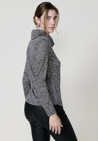 Grau, strukturiertes Langarmoberteil mit einem Wasserfallausschnitt, kombiniert mit schwarzen, eng anliegenden Jeans. Der Stoff wirkt weich und leicht dehnbar.