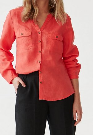 Femme portant une chemise corail boutonnée avec poches poitrine et un pantalon noir, une main dans la poche, sur un fond clair uni.
