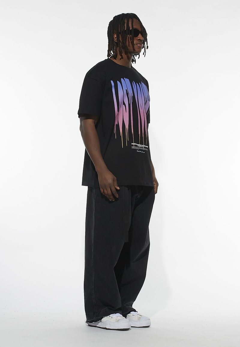 T-shirt nera oversize con design testuale in gradiente "LAST" in blu e rosa. Abbinata a pantaloni neri larghi e sneakers bianche.