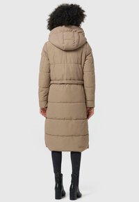 Lange, gevoerde jas in beige met een capuchon en een getailleerde taille. Heeft een gewatteerde textuur en een logo label nabij de zoom. Gecombineerd met zwarte broek en laarzen.