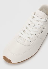 Adidași din piele albă cu șireturi albe, vârf rotunjit și talpă din cauciuc bej. Prezintă inscripția "TOMMY HILFIGER" embosată pe lateral. Textură netedă.