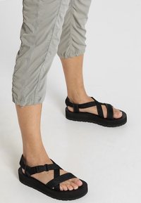 Nogi w jasnoszarych, marszczonych spodniach i czarnych platformach na grubej podeszwie z paskami i klamrami na białym tle.