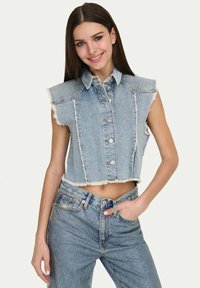 Gilet in denim cropped con chiusura a bottoni, colore azzurro chiaro, bordi sfilacciati e design senza maniche; indossato con jeans a vita alta abbinati.