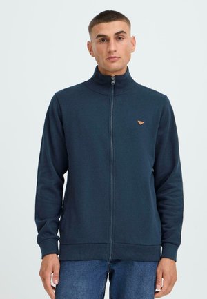 Sweatshirt zippé bleu marine avec col haut, poignets et ourlet côtelés. Présente un petit logo marron sur la poitrine, fabriqué en tissu doux.