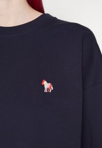 Granatowa bluza z prążkowanym wykończeniem przy dekolcie. Na lewej stronie klatki piersiowej znajduje się niewielki, wielokolorowy haftowany koń. Miękka tekstura materiału.