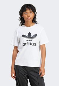 Camiseta de algodón blanca con un logo negro de Adidas y un diseño de tres rayas. Mangas cortas y un corte relajado. Combinada con pantalones negros.