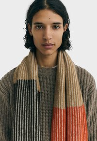 Knitted scarf featuring vertical stripes in tan, black, cream, and orange, draped over a textured brown sweater, with a soft, warm texture.
