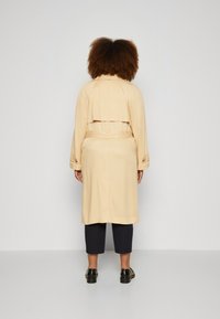 Trench beige con un diseño acortado, cintura ceñida con cinturón y mangas abotonadas, combinado con pantalones navy cortos y zapatos negros.