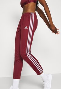 Bordeauxfarbene Adidas-Sporthose aus weichem Material, mit weißen Seitenstreifen, Druckknopfdetails und einer körpernahen Passform mit Knöchelabschlüssen.