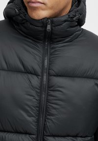 Close-up van een persoon in een zwarte pufferjack met een ritssluiting aan de voorkant en verstelbare koorden op de capuchon.