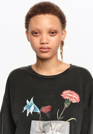 Persona joven con cabello corto y rizado que lleva una camisa negra con estampado de flores azules y rojas y un pendiente largo de borla dorada en una oreja.