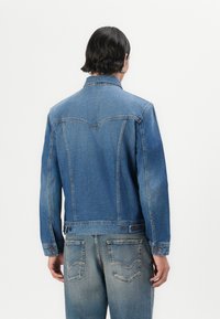 En person med mellomlangt mørkt hår har på seg en denimjakke, vendt bort fra kameraet, mot en enkel hvit bakgrunn. Jakken har klassisk stil.