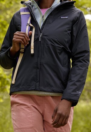 Personne portant une veste noire Patagonia, un polaire bleu clair, un pantalon rose, et portant une sangle de sac à dos violette à l'extérieur.