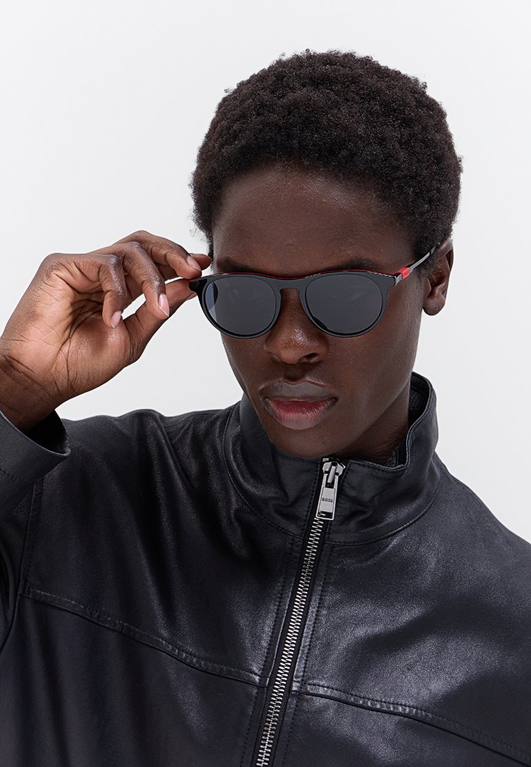 Occhiali da sole aviator neri con lenti scure, montatura nera sottile e dettagli rossi, indossati da una persona in giacca di pelle nera.