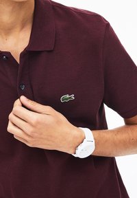Polo bordeaux en tissu texturé, avec un logo crocodile vert brodé, deux boutons noirs et un col classique.