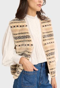 Beige Kunstpelz-Weste mit filigranen schwarzen und braunen Musterdetails, ausgestattet mit einem Knopfverschluss und zwei Seitentaschen. Getragen über einer weißen Bluse.