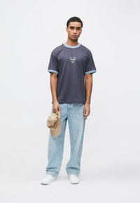 Νέος άνδρας που φοράει σκούρο γκρι μπλουζάκι με γαλάζιο περιθώριο, γαλάζια τζιν παντελόνια, λευκά αθλητικά παπούτσια, κρατώντας ένα μπεζ καπέλο στο αριστερό του χέρι.