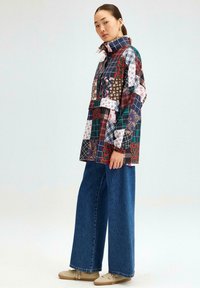 Multikleurige patchwork shirt met hoge kraag, bloemen- en geometrische patronen, gecombineerd met wijde denim broek en beige schoenen.