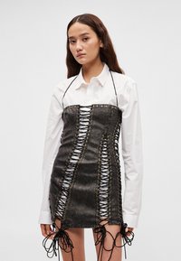 Crna haljina od umjetne kože s detaljima za vezanje, s otvorma uz bočne strane i podesivim vezicama na donjem rubu. Nošena preko bijele košulje.