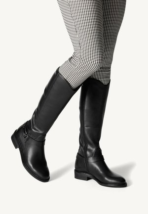 Bottes en cuir noires montantes jusqu'aux genoux avec des talons bas, portées sur un pantalon à motif pied-de-poule noir et blanc, jambes croisées.