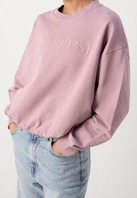Světle růžová mikina s kulatým výstřihem a vyšitým logem "Marc O’Polo". Má spadlé ramena a žebrované manžety, kombinováno se světle modrými džíny.