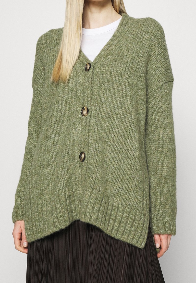 Cardigan en maille lâche vert olive avec trois boutons marbrés porté sur un haut blanc, associé à une jupe plissée noire.