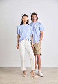 Hellblaue T-Shirts mit floralem Design auf der linken Brust, kombiniert mit weißen, hochgeschnittenen Hosen und beigen Shorts. Keilabsatzschuhe und Sneakers werden getragen.