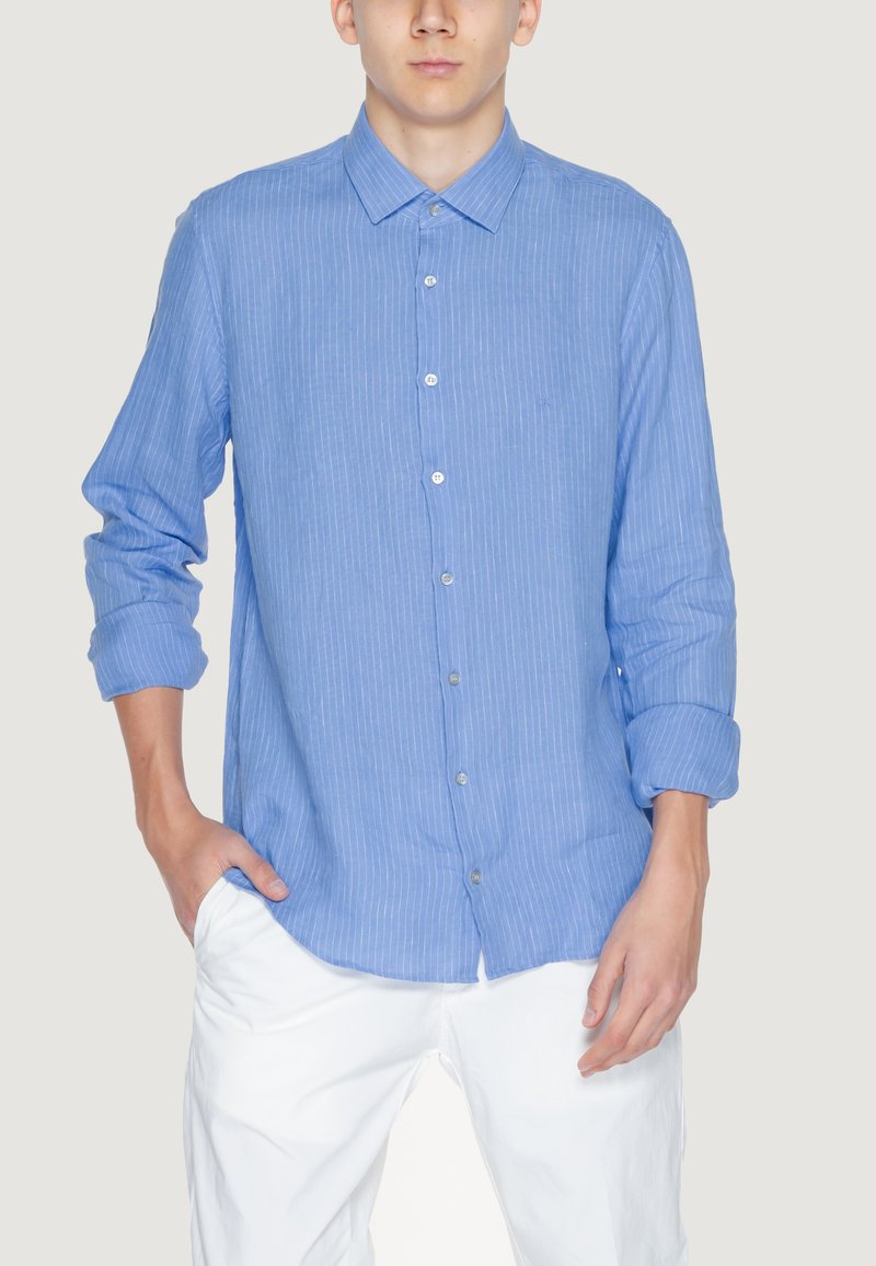 Uomo che indossa una camicia a maniche lunghe, blu chiaro, con bottoni, a strisce verticali sottili bianche e pantaloni bianchi, in piedi con una mano in tasca.