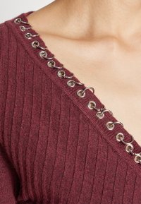 Gros plan sur un haut en maille côtelée bordeaux avec un décolleté diagonal orné d'anneaux et de crochets en métal argenté.