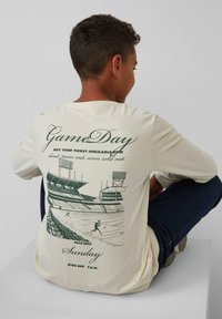 Cremefarbenes Langarmshirt mit grünen Druckgrafiken eines Stadions und eines Spielers, mit Text, der Informationen zu Spieltag und Tickets enthält.