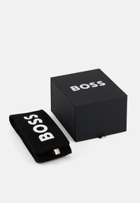 Must plaat, mille valge "BOSS" logoga, sisaldab vastavat musta kangast aksessuaari koos logoga. Sile tekstuur, ristkülikukujuline kuju, minimaalne disain.
