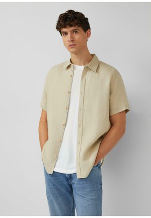 Junger Mann mit lockigem Haar, der ein beige kurzärmeliges Hemd über einem weißen T-Shirt und blauen Jeans trägt, steht mit den Händen in den Taschen.