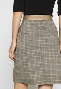 Jupe à carreaux beige et noir avec une large ceinture beige lisse et deux poches arrière. Le tissu a une texture subtile.